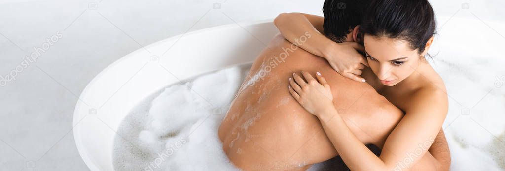 Beautiful naked woman hugging boyfriend in bathtub with foam on grey background, panoramic shot
