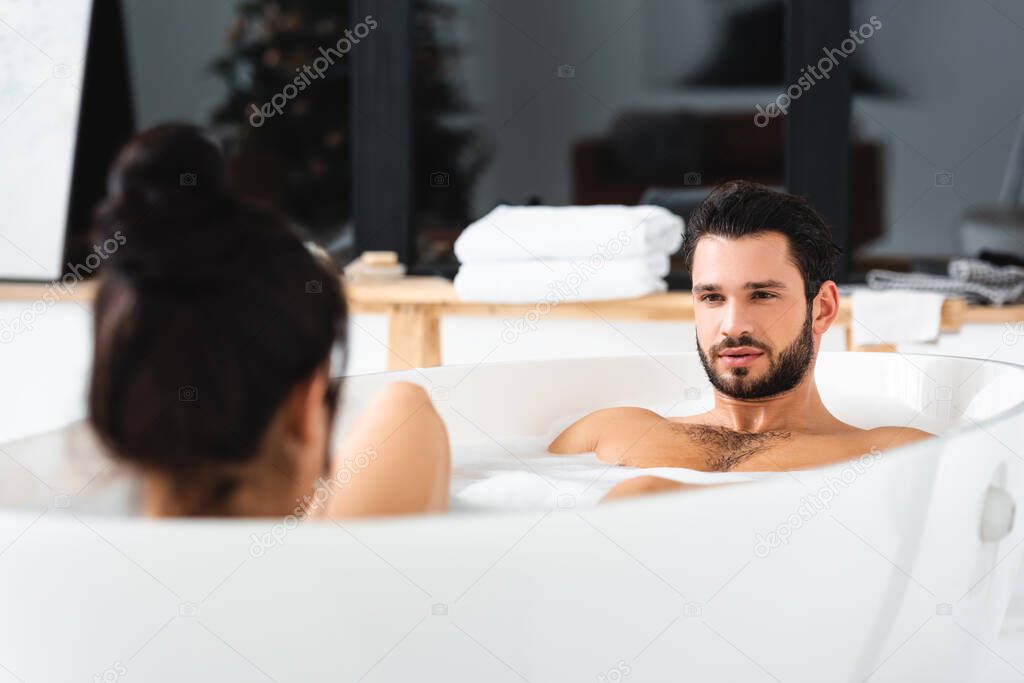 Selective focus of handsome man looking at girlfriend in bathtub with soapsuds
