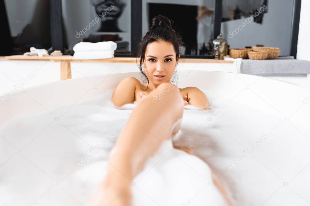 Selective focus of sensual woman looking at camera while taking bath with foam at home