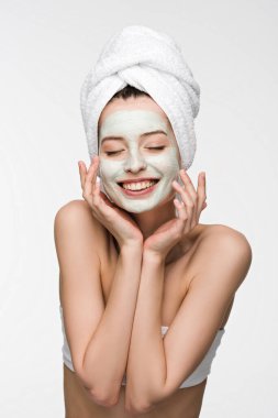 cheerful girl with nourishing facial mask and towel on head touching face with closed eyes isolated on white