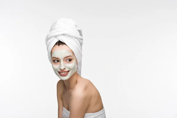 cheerful girl with facial nourishing mask and towel on head looking away isolated on white