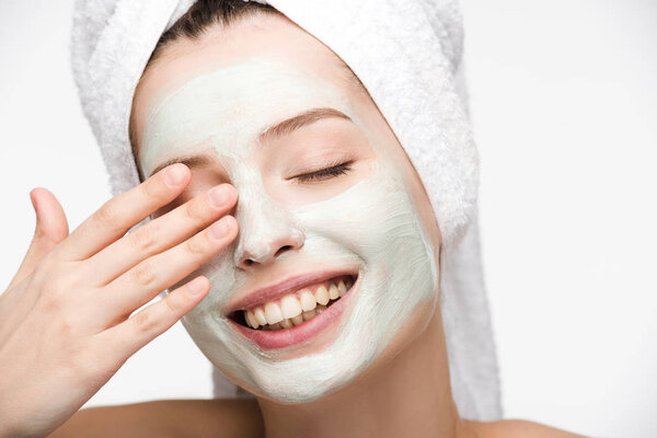 happy girl with facial nourishing mask and towel on head touching eye isolated on white