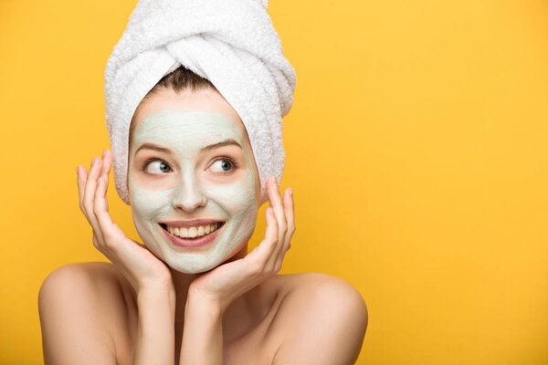 cheerful girl with nourishing facial mask and towel on head looking away isolated on yellow