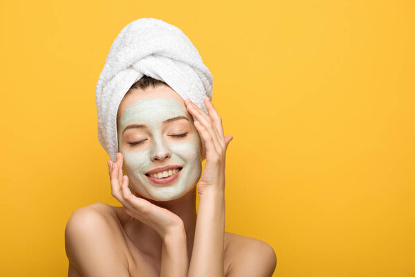 happy girl with nourishing facial mask and towel on head touching face with closed eyes isolated on yellow