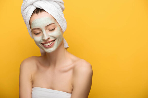 smiling girl with nourishing facial mask standing with closed eyes isolated on yellow