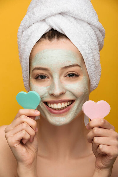 cheerful girl with nourishing mask on face holding heart-shaped cosmetic sponges isolated on yellow