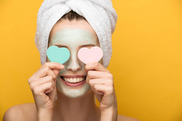 smiling girl with nourishing facial mask covering eyes with heart-shaped cosmetic sponges on yellow background 