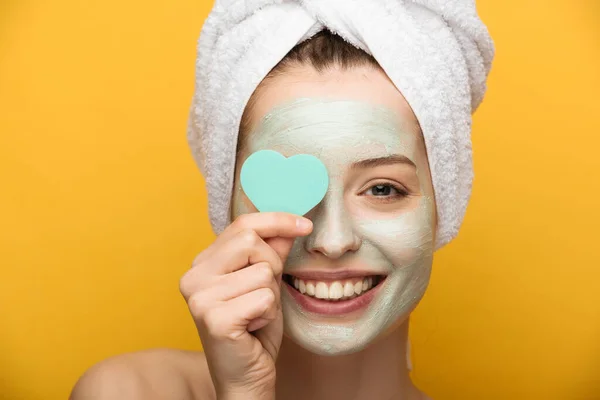 happy girl with nourishing mask on face covering eyes with heart-shaped cosmetic sponge on yellow background