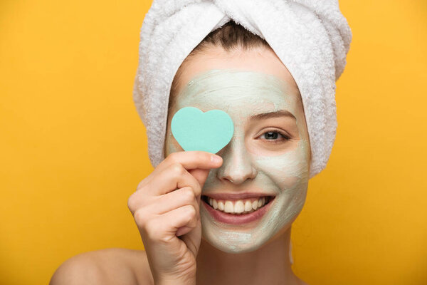happy girl with nourishing mask on face covering eyes with heart-shaped cosmetic sponge on yellow background