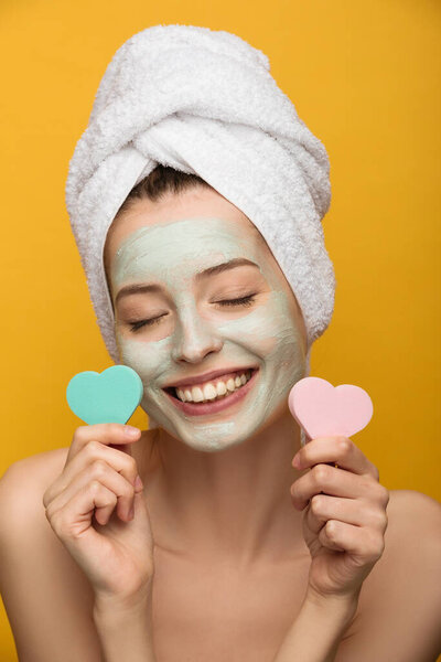 cheerful girl with nourishing facial mask holding heart-shaped cosmetic sponges with closed eyes isolated on yellow