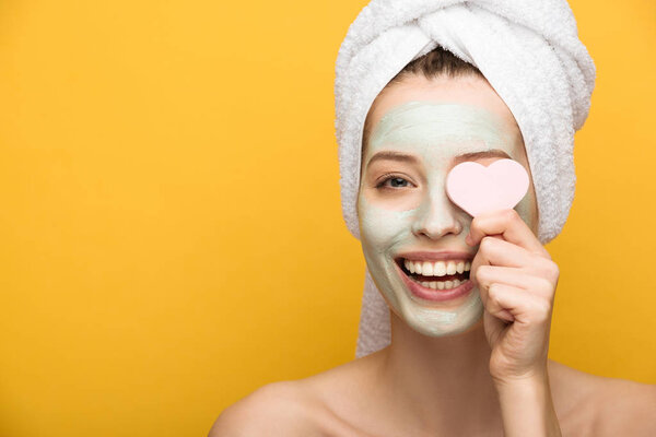 cheerful girl with nourishing facial mask covering eyes with heart-shaped cosmetic sponge isolated on yellow