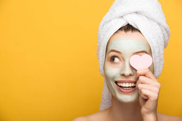 cheerful girl with nourishing facial mask covering eye with heart-shaped cosmetic sponge on yellow background