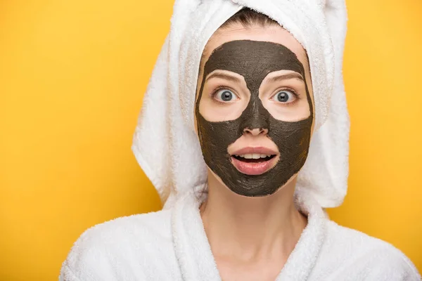 shocked girl with facial clay mask and towel on head looking at camera on yellow background