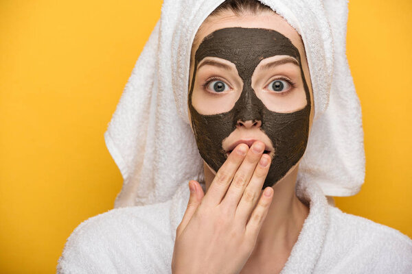в шоке девушка с лицом глиняная маска и полотенце на голове покрывающий рот с рукой изолированы на желтый
