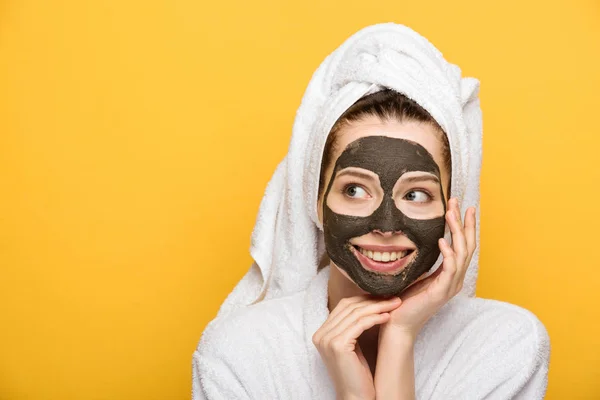 happy girl with facial clay mask and towel on head looking away isolated on yellow