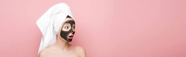 panoramic shot of shocked girl with facial clay mask and towel on head looking away isolated on pink