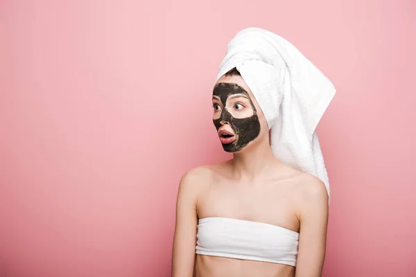 shocked girl with facial clay mask looking away while standing with open mouth on pink background