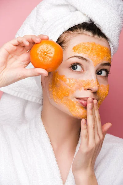 魅力的な女の子とともに柑橘系の顔のマスクはピンクに隔離されたみかんを保持しながら唇に触れる — ストック写真