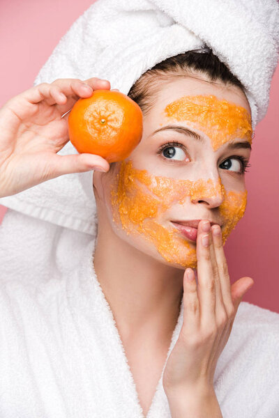 attractive girl with citrus facial mask touching lips while holding tangerine isolated on pink