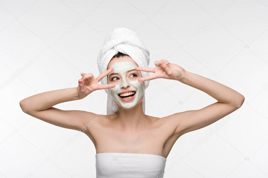 cheerful girl with facial nourishing mask and towel on head showing victory gestures isolated on white