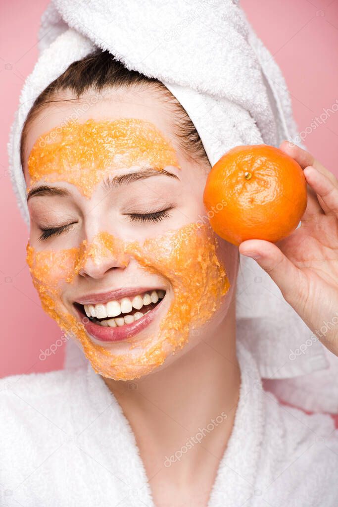 smiling girl with citrus facial mask holding tangerine with closed eyes isolated on pink