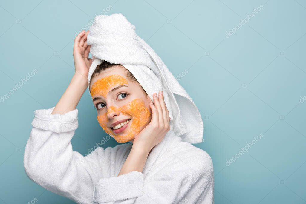smiling girl with citrus facial mask touching face and towel on head while looking away on blue background