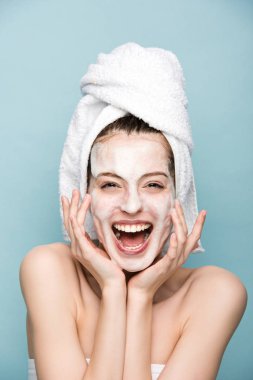 excited girl with moisturizing facial mask laughing at camera isolated on blue clipart