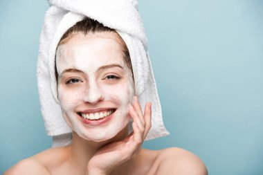 cheerful girl with moisturizing facial mask touching face while looking at camera isolated on blue clipart