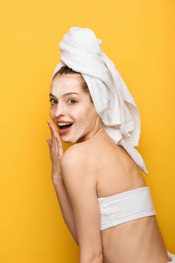 surprised girl with moisturizing facial mask holding hand near mouth and looking at camera isolated on yellow  clipart