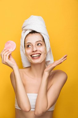 Yüzünde nemlendirici maske olan mutlu bir kız sarıya izole edilmiş kozmetik süngerine bakıyor.