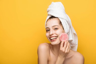 happy girl with moisturizing facial mask looking at cosmetic sponge isolated on yellow clipart