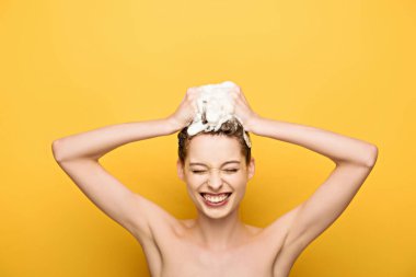cheerful girl smiling with closed eyes while washing hair on yellow background clipart