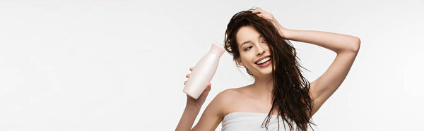 panoramic shot of smiling girl touching clean hair while holding shampoo isolated on white