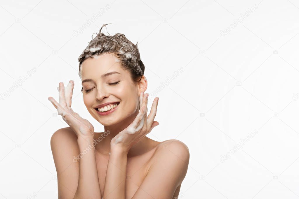 smiling girl with soapy hair and closed eyes holding hands near face isolated on white
