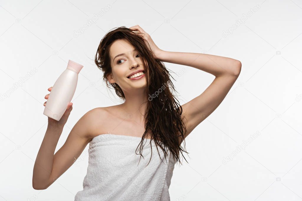 smiling girl touching clean hair while holding shampoo isolated on white