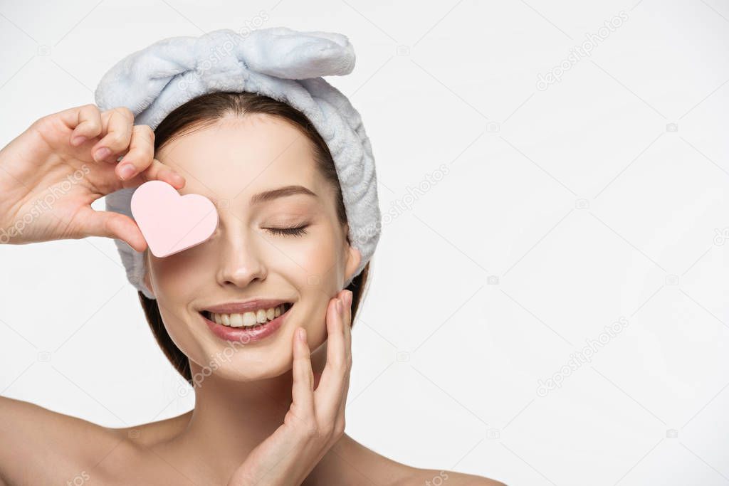 happy girl covering eye with heart-shaped cosmetic sponge while touching face isolated on white
