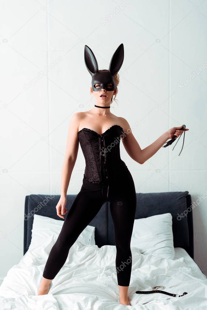 sexy woman in bunny mask holding flogging whip while standing on bed 