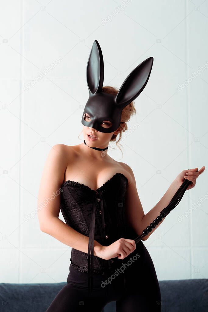 dominant young woman in bunny mask and corset holding flogging whip and looking away 