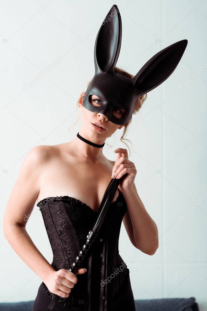 dominant young woman in bunny mask and corset holding flogging whip and looking at camera 