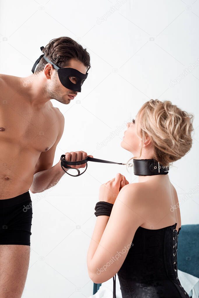 man in eye mask in holding bdsm leash and looking at submissive woman 