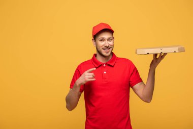 Kırmızı üniformalı mutlu teslimatçı, sarı üzerine izole edilmiş pizza kutusunu işaret ediyor.