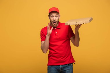 Kırmızı üniformalı mutlu teslimatçı elinde pizza kutusu, ağzı açık, sarıda izole edilmiş.