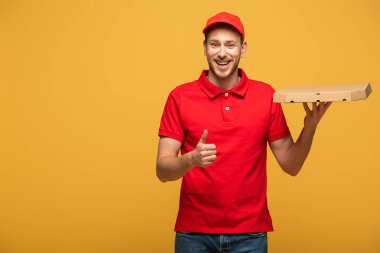 Kırmızı üniformalı mutlu teslimatçı elinde pizza kutusuyla sarıda izole bir şekilde baş parmağını kaldırıyor.