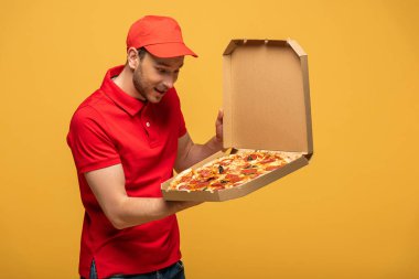 Kırmızı üniformalı mutlu teslimatçı, sarı izole edilmiş lezzetli pizzayla dolu kutuya bakıyor.