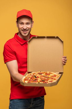 Kırmızı üniformalı mutlu teslimatçı, sarı üzerine izole edilmiş lezzetli pizzayla kutusunu gösteriyor.