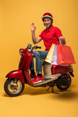 Kırmızı üniformalı gülümseyen teslimatçı, sarı arka planda scooter 'a el sallıyor.