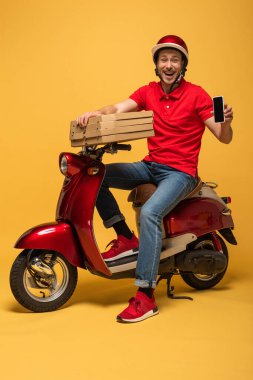 Kırmızı üniformalı mutlu teslimatçı elinde pizza kutuları ve sarı arka planda scooter 'da boş ekranlı akıllı telefon tutuyor.
