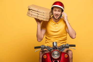 Sarı üniformalı gülümseyen teslimatçı elinde pizza kutularıyla scooter sürüyor ve sarı renkte izole edilmiş akıllı telefondan konuşuyor.