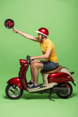 Sarı üniformalı teslimatçının elinde tavayla motosikletinin üstünde yeşil bir fiyonk tutarken yan görüntüsü.
