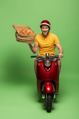 Sarı üniformalı teslimatçı motosikletli, yeşil yolda pizza dağıtıyor.